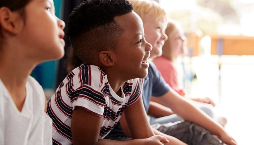 Group Of Elementary School Pupils Sitting On Floor Listening To Teacher